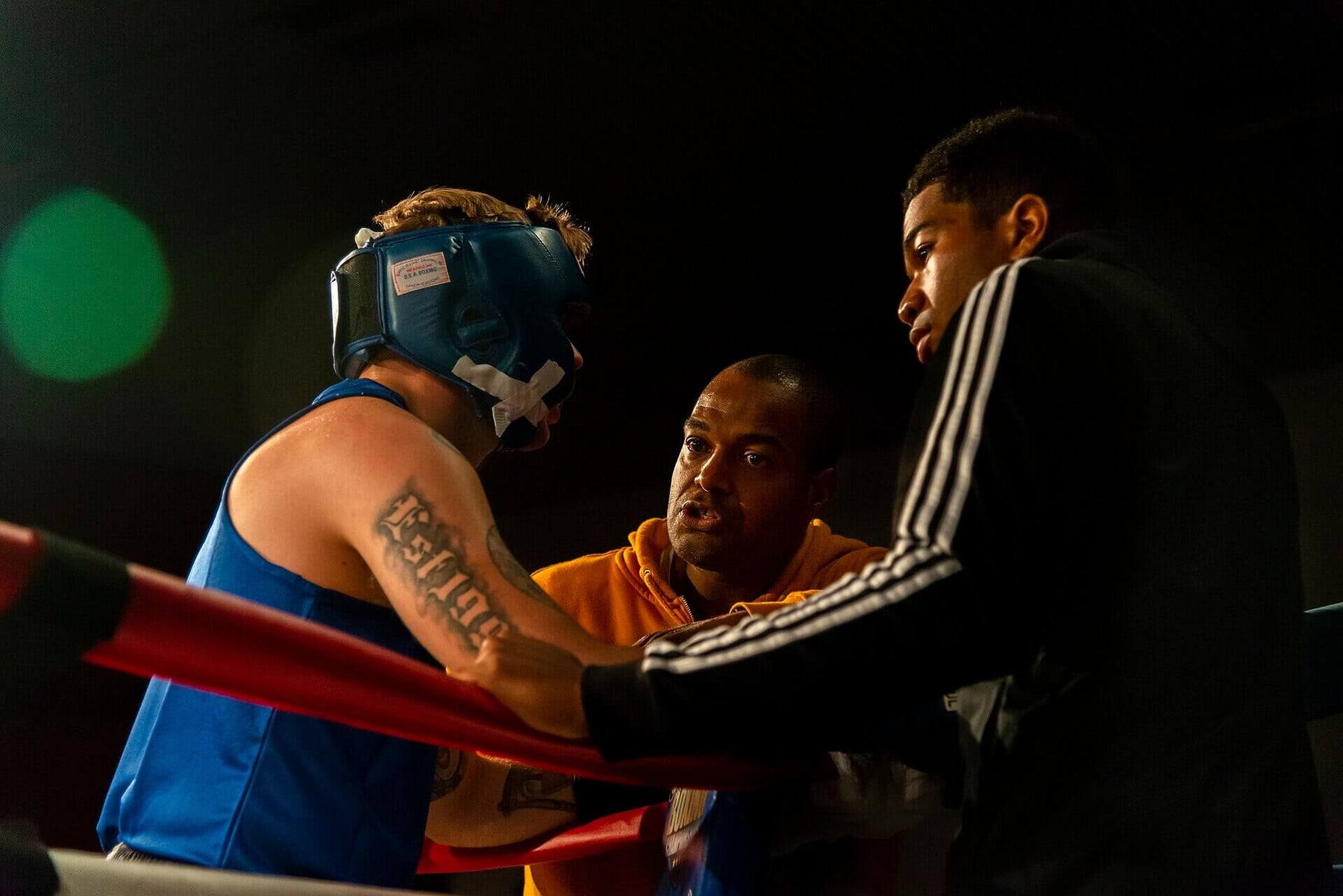 A boxing coach speaks closely to an amateur boxer in blue headgear between rounds, while another corner team member leans over the red ring ropes.