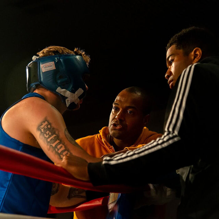 A boxing coach speaks closely to an amateur boxer in blue headgear between rounds, while another corner team member leans over the red ring ropes.