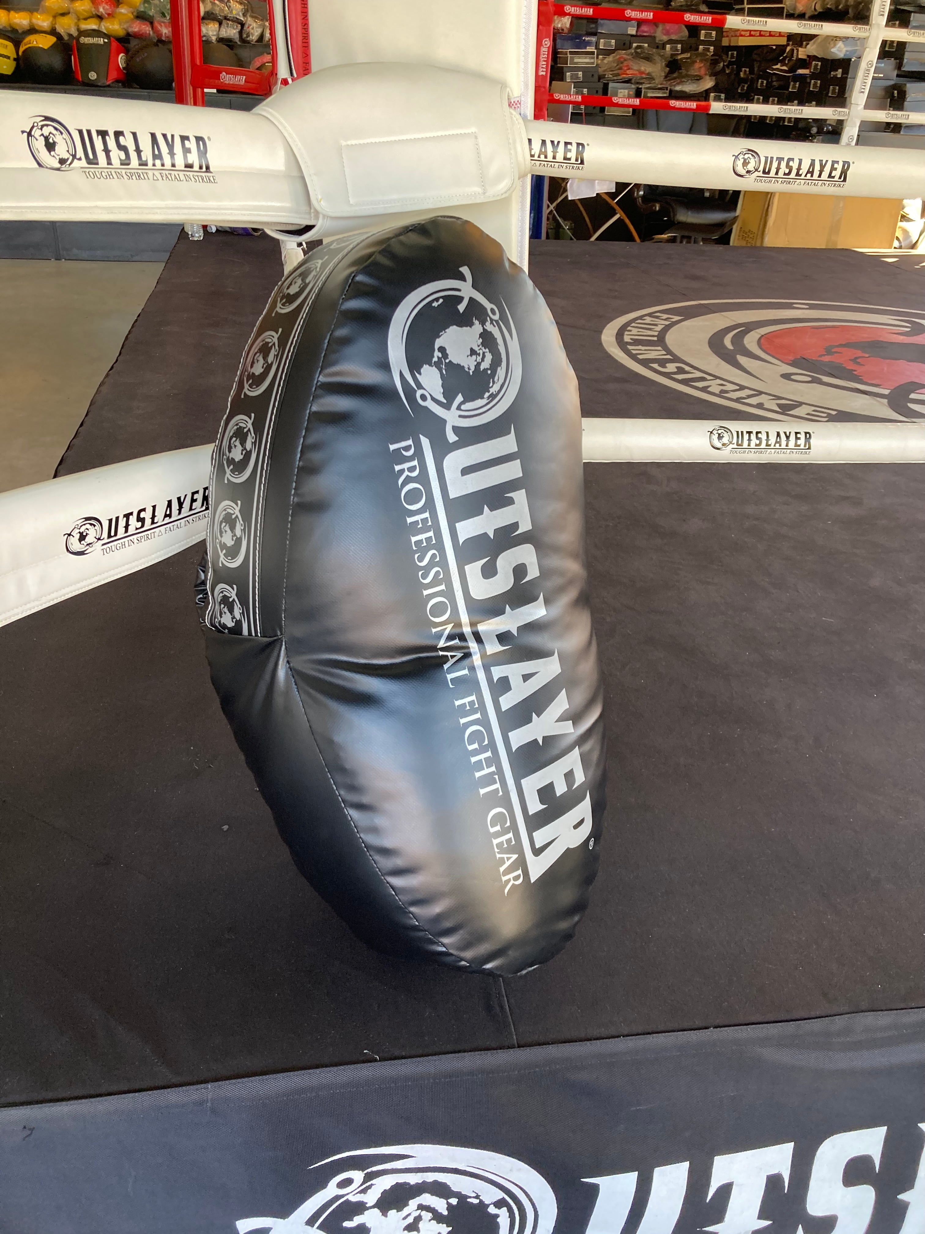 A black weighted kicking shield/pad with white 'OUTSLAYER' branding, positioned inside a boxing ring on a dark mat.