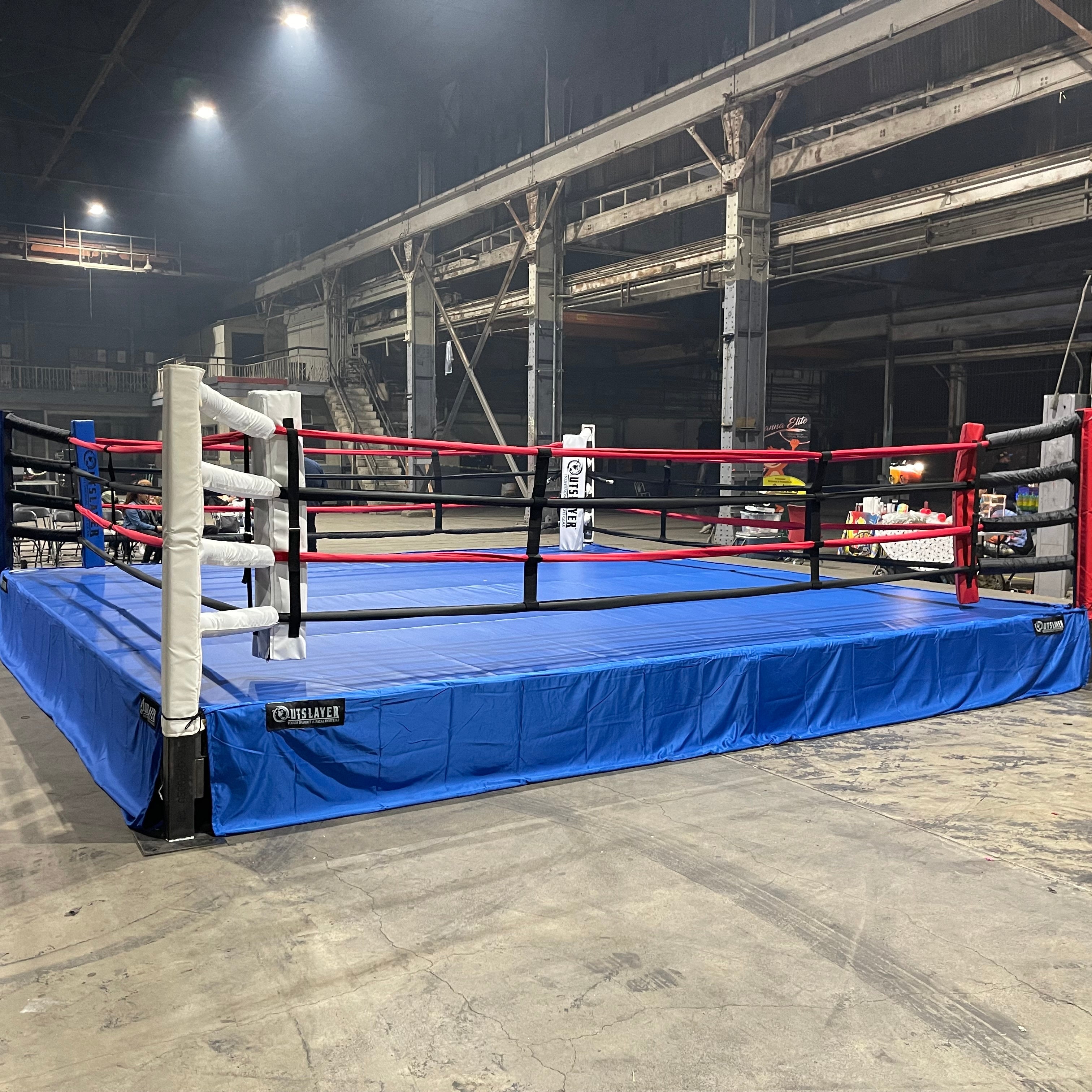 A professional boxing ring with a blue flooring, black ropes, and red and white padding. The ring is set in an indoor location with a dark background.