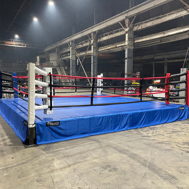 A professional boxing ring with a blue flooring, black ropes, and red and white padding. The ring is set in an indoor location with a dark background.